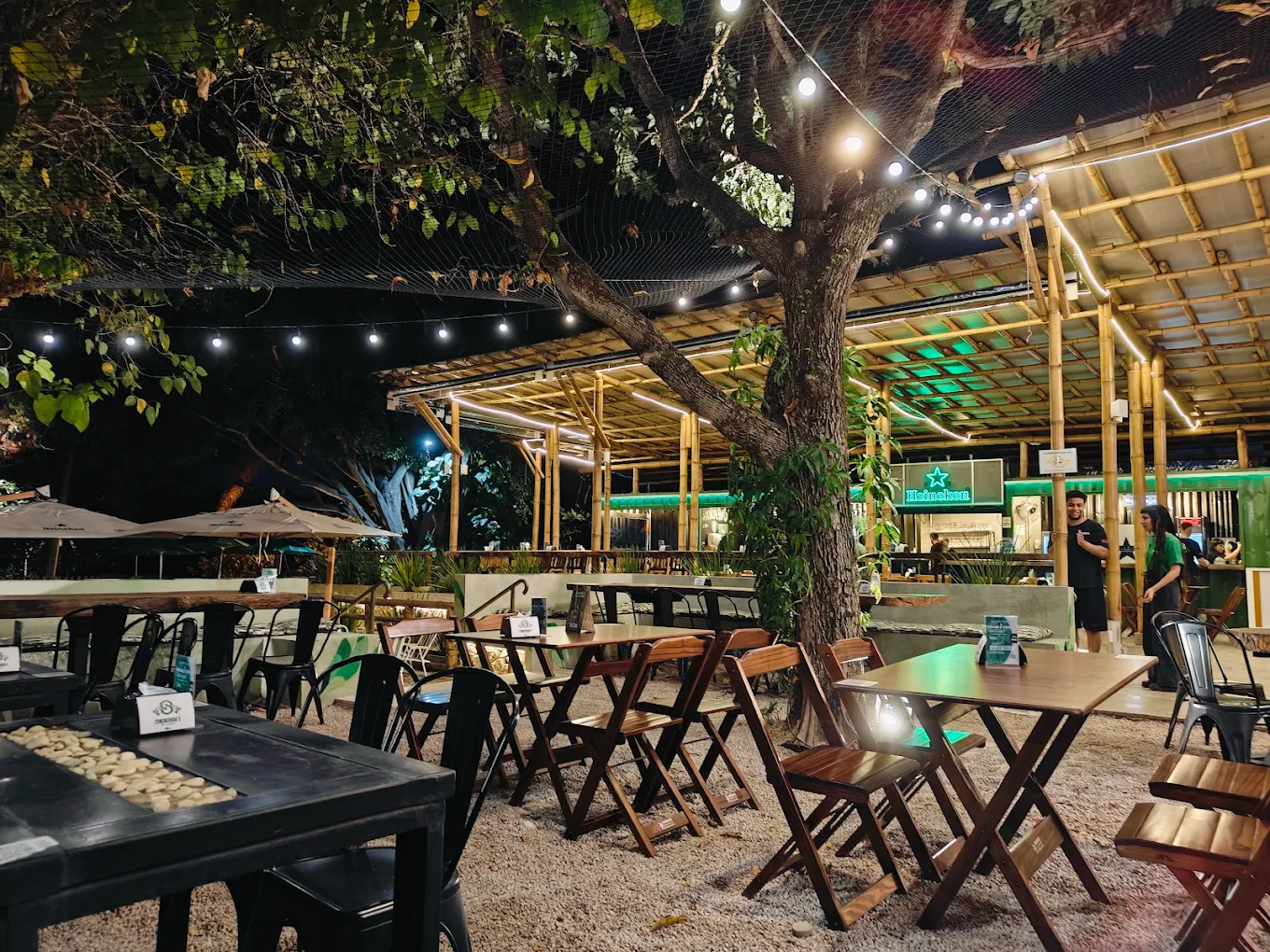 Mesas de madeira sob a árvore central do Quintal à noite, com luzes penduradas e o salão de bambu ao fundo