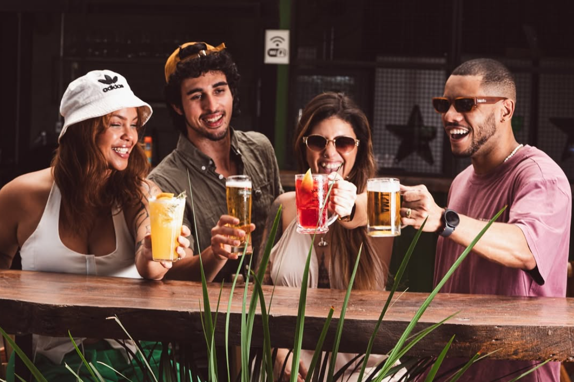Quatro amigos sorrindo e brindando com chope e drinks em mesa alta de madeira do Quintal