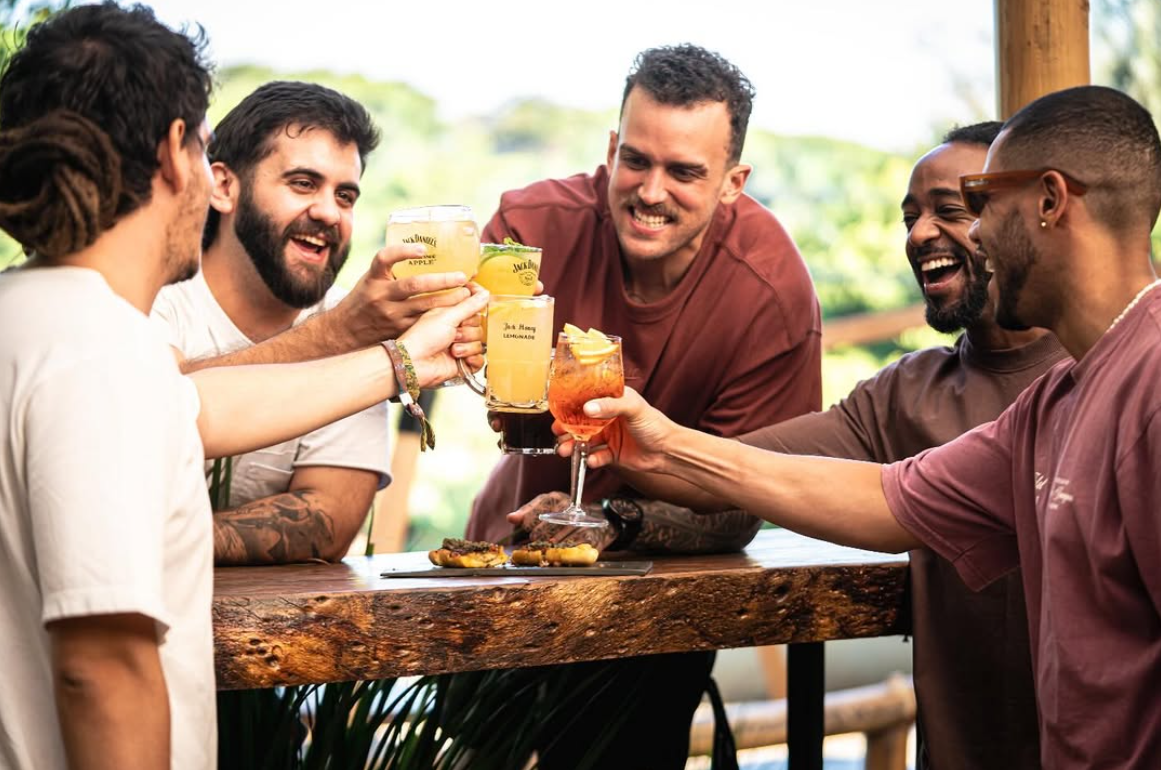 Grupo de amigos rindo e brindando com drinks e chope sobre a mesa de madeira maciça do Quintal, com verde ao fundo