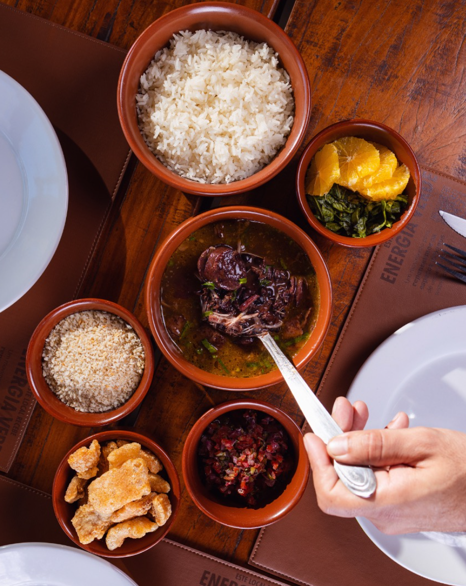 Mesa servida com feijoada completa, acompanhamentos e chope no Quintal
